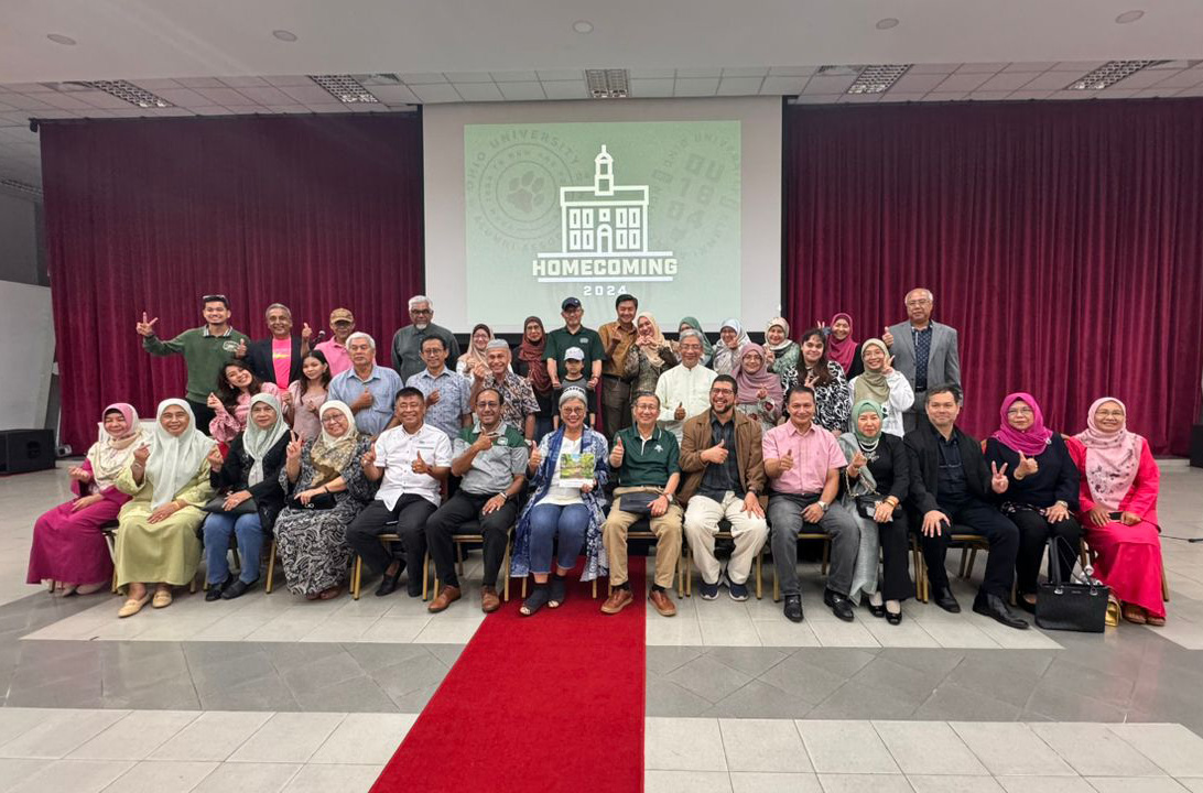 group photo of nearly 40 people celebrating OHIO's Homecoming 2024, smiling and posing in front of a maroon curtain