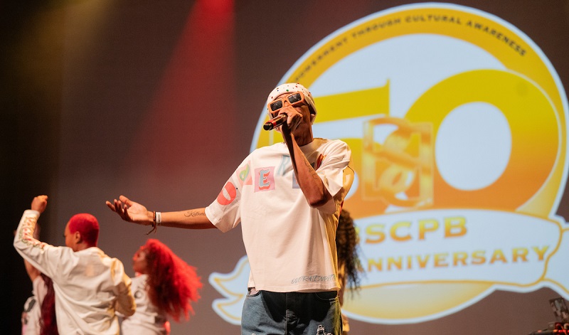 A performer is shown on stage in front of the BSCPB Anniversary Logo