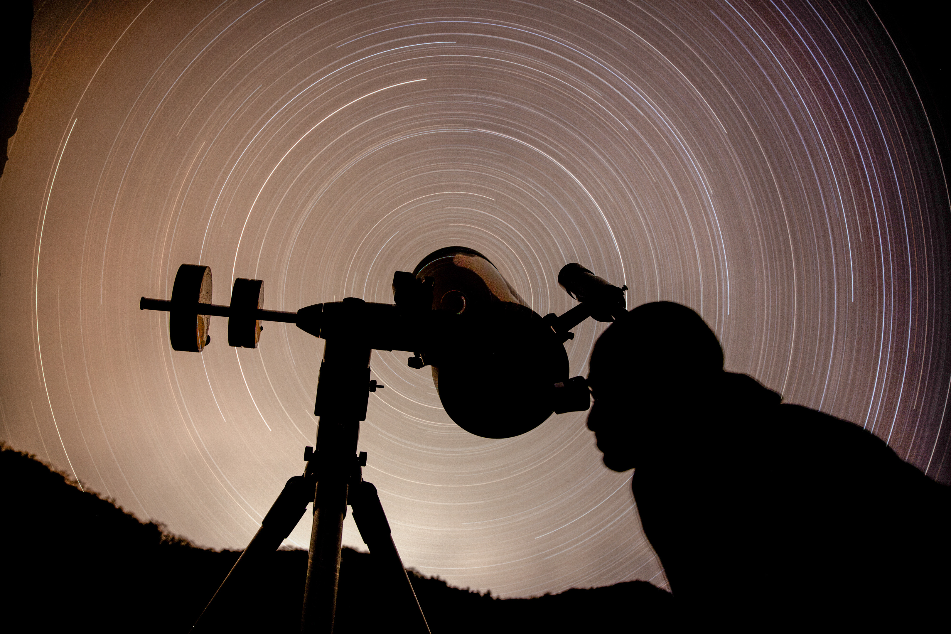 A silhouette of a telescope against a long-exposure blur of a starry sky.