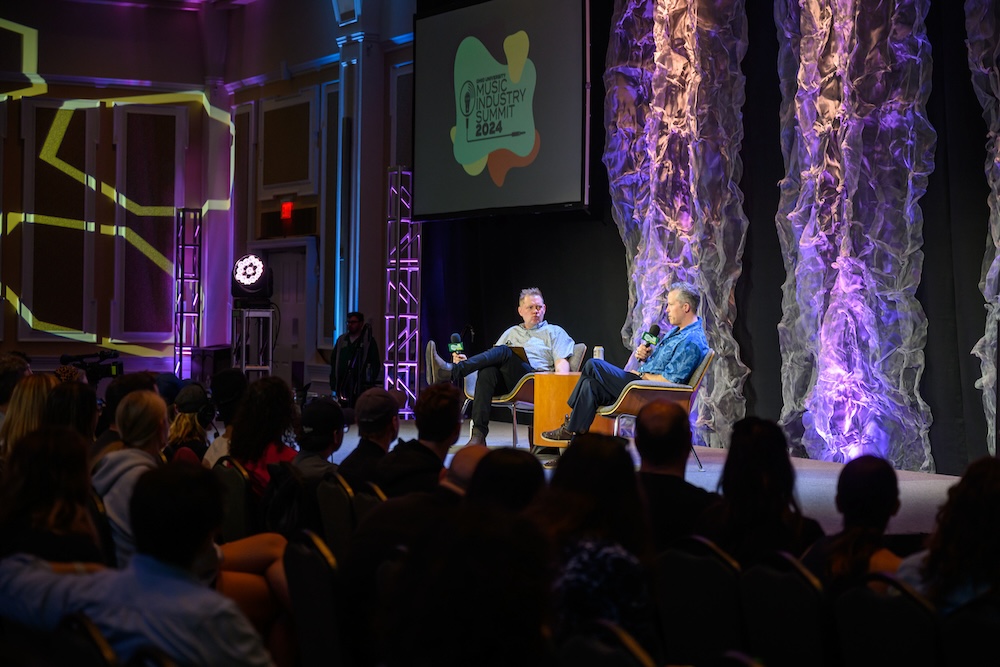 Two men sit on a stage, speaking to each other