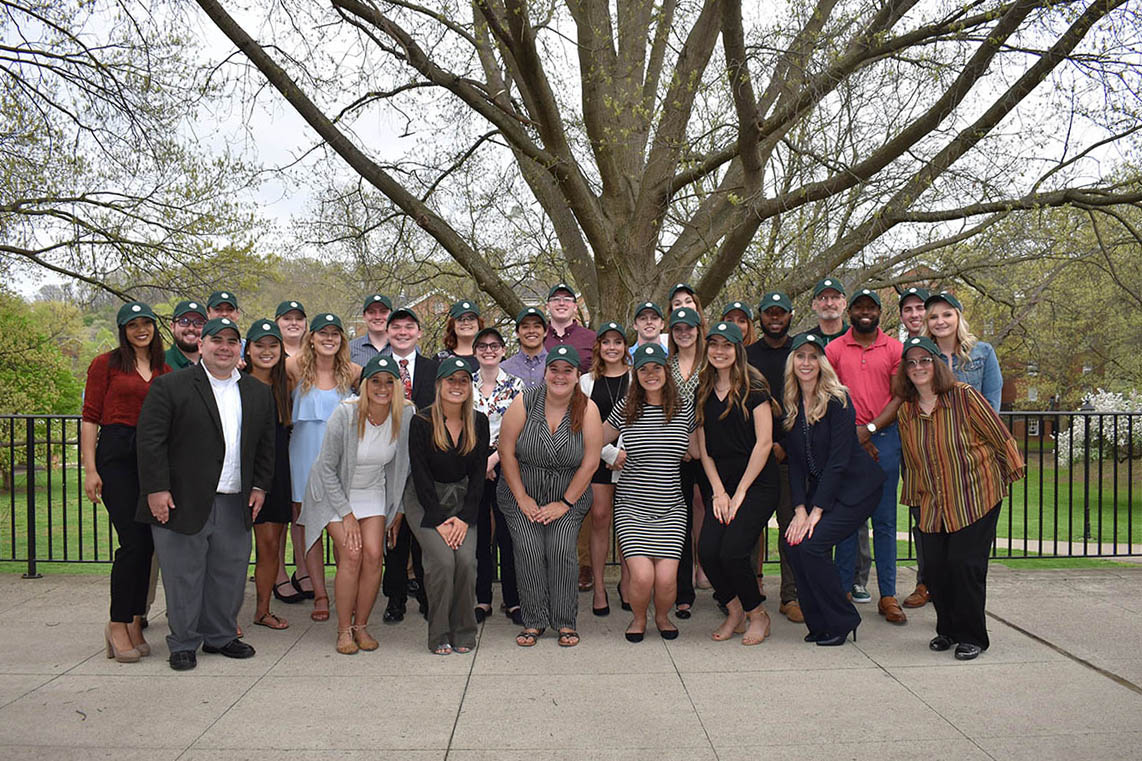 a group of about 30 students and faculty advisors pose wearing green ballcaps under a tree with spring leaves budding