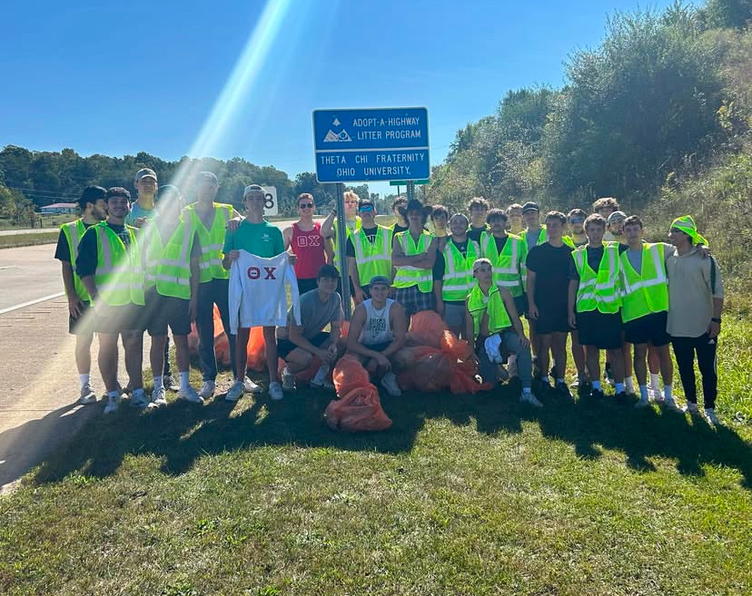 a group of about two dozen young men wearing safety vests pose next to their "adopt a highway" sign with trash bags full of litter