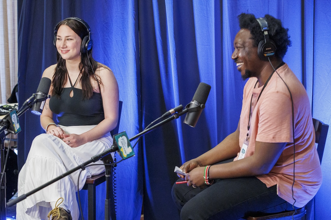 Two people sit in front of microphones in front of a blue curtain backdrop, smiling