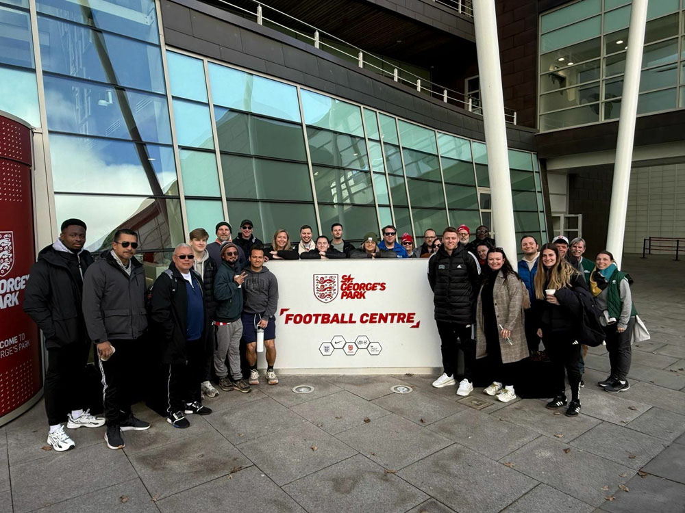 A group poses for a photo while holding the St. George’s Park Football Centre sign.