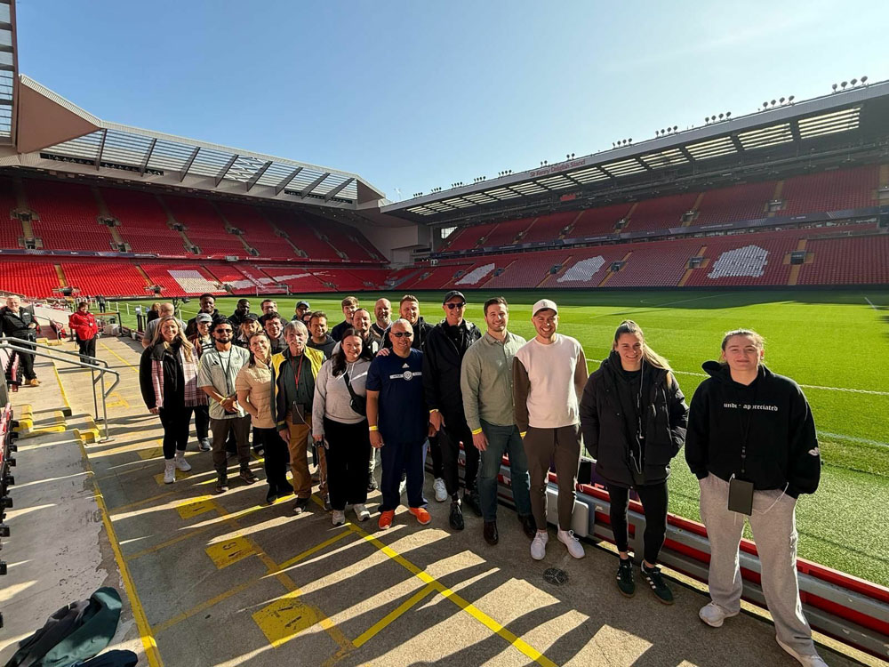 A group of people pose for a photo inside a large, empty soccer stadium on a sunny day.