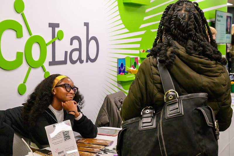 An OHIO student sits at a table at the CoLab and explains her products to another student