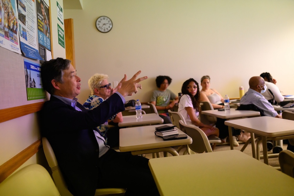 A man sits in a classroom, gesturing while responding to a presenter