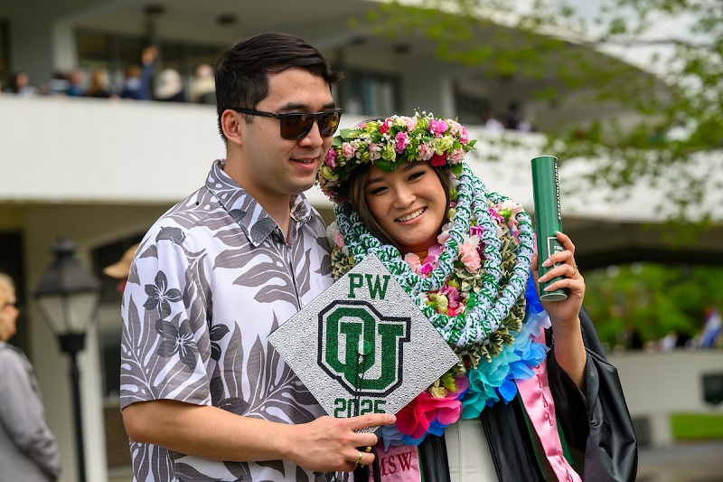 An OHIO graduate wearing a colorful items over her cap and gown poses for a photo with an individual outside of the Convocation Center after Spring Commencement