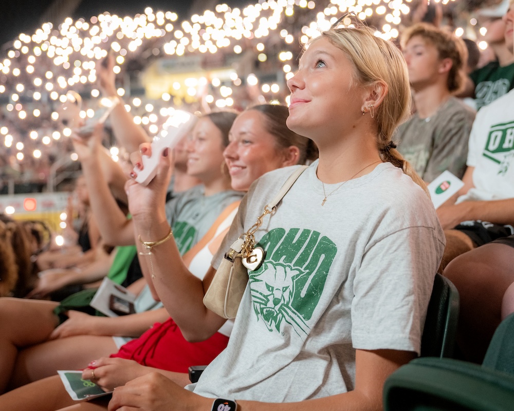 Sitting in a large crowd doing the same, a student holds up her phone's flashlight