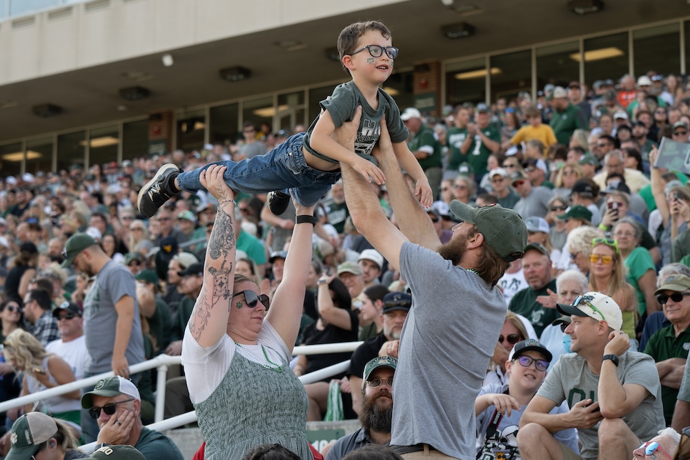 Two adults hold up a child in OHIO gear in the stands of the Homecoming football game