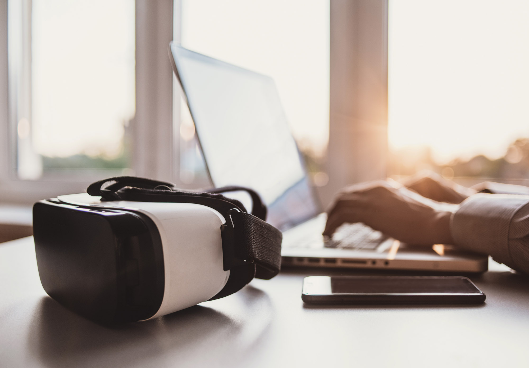 Virtual reality headset on a desk with someone on a laptop behind it.