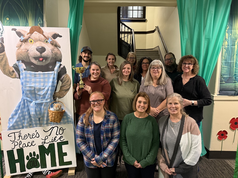 OHIO employees pose with their trophy in the Business Service Center, which has a sign that features a Bobcat and the words There's No Place Like Home