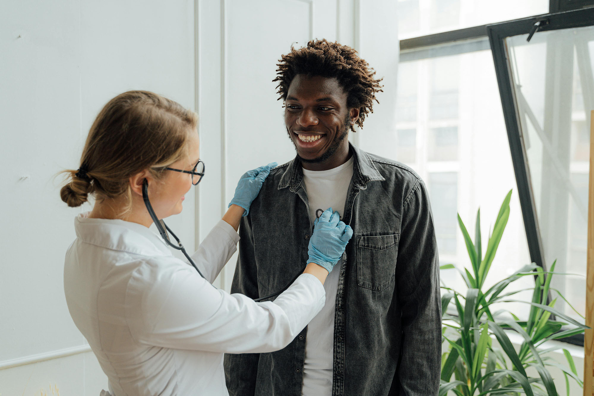 a female doctor wearing gloves and a white coat uses a stethoscope to listen to a smiling patient’s chest during an exam in a brightly lit room