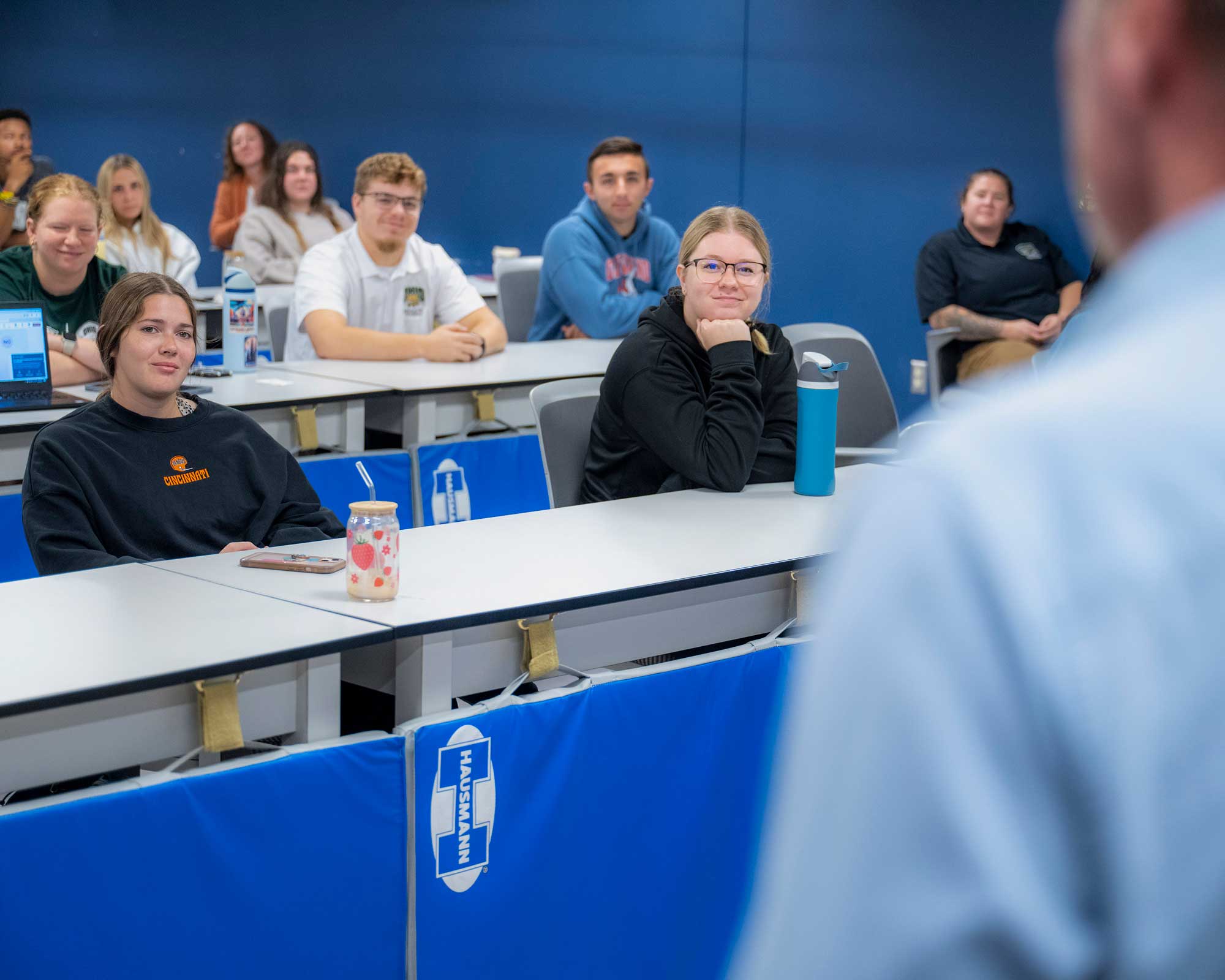 a group of students sitting at desks listen as a speaker in the foreground addresses them