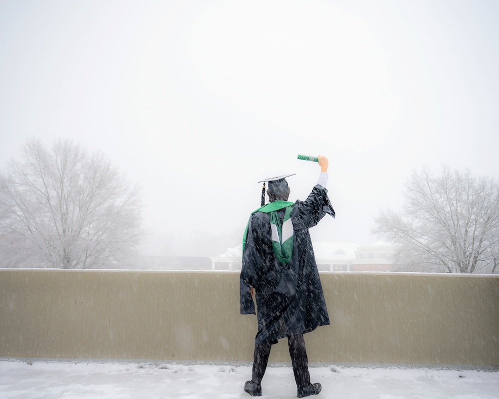 Pictured from behind, a graduate in cap and gown holds up a diploma outside the Convo, snow falling around him