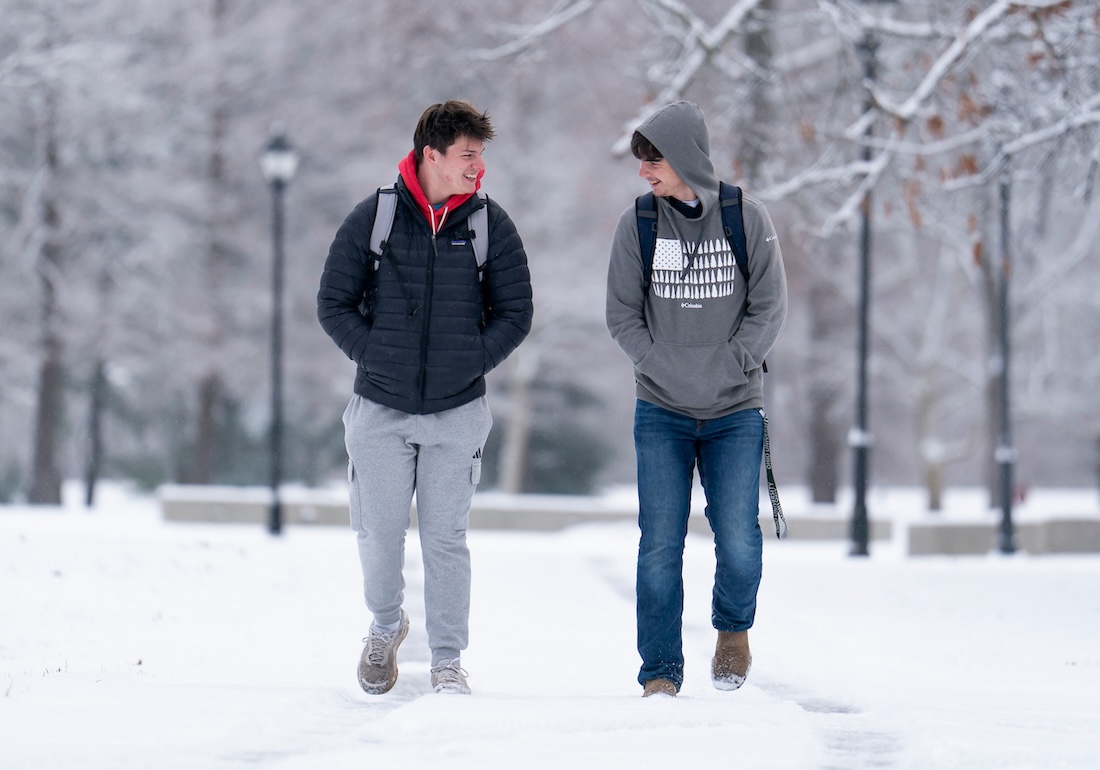 Two students walk through the snow, facing each other and laughing