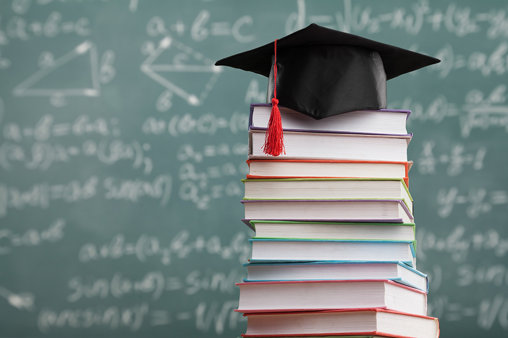 A graduation cap perched atop a pile of books in front of a green chalkboard with equations written on it
