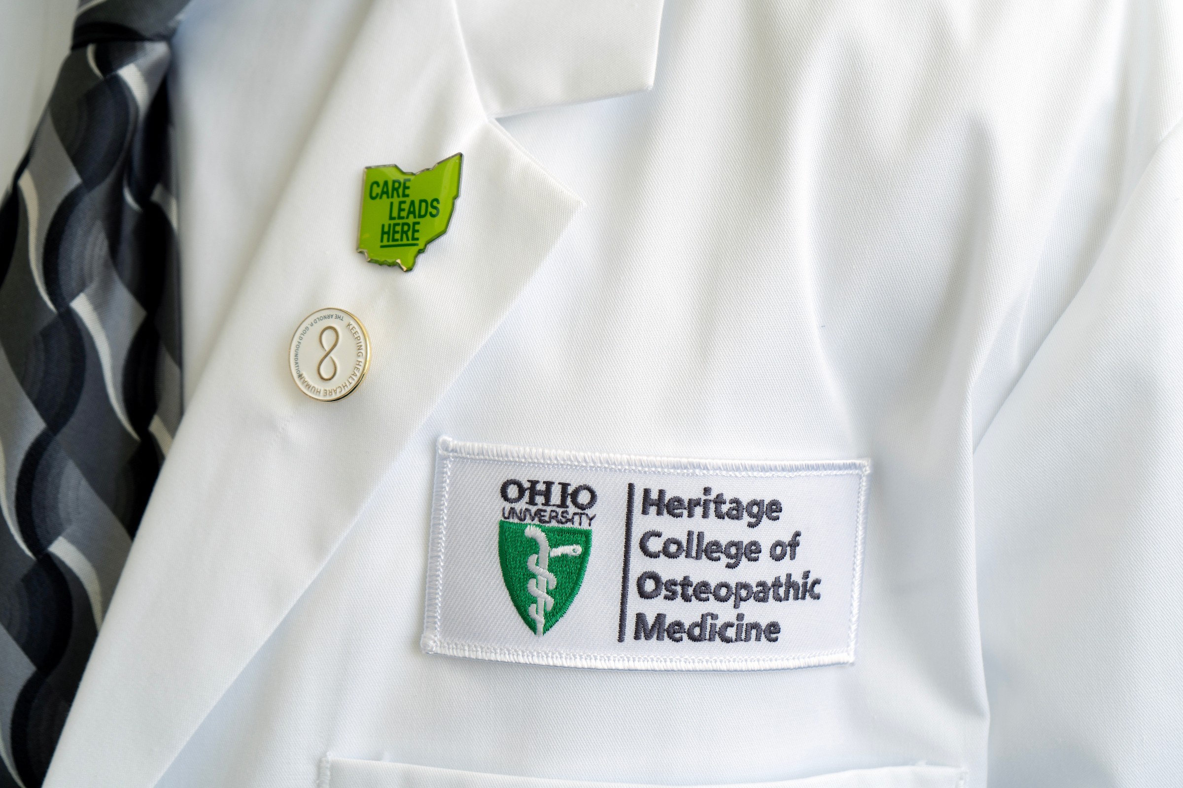 The Heritage College of Osteopathic Medicine logo is shown on a white coat, worn by a doctor. The coat also has a pin that says Care Leads Here
