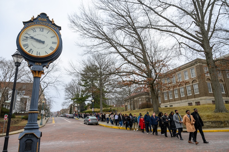 A group of people march arm-in-arm past a large clock on OHIO's campus
