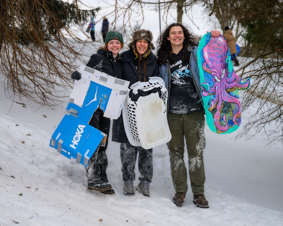 Three students stand side-by-side in the snow, smiling and holding up a flattened shoe box, a destroyed laundry basket, and a blue and purple sled