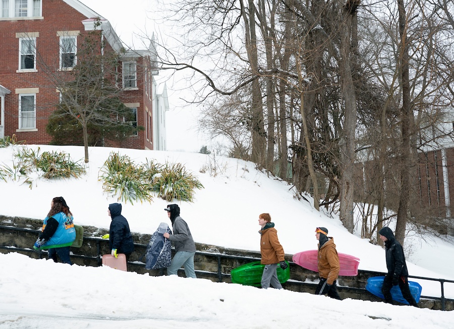A group of students walk up stairs in the snow, all carrying sleds and dressed for cold weather