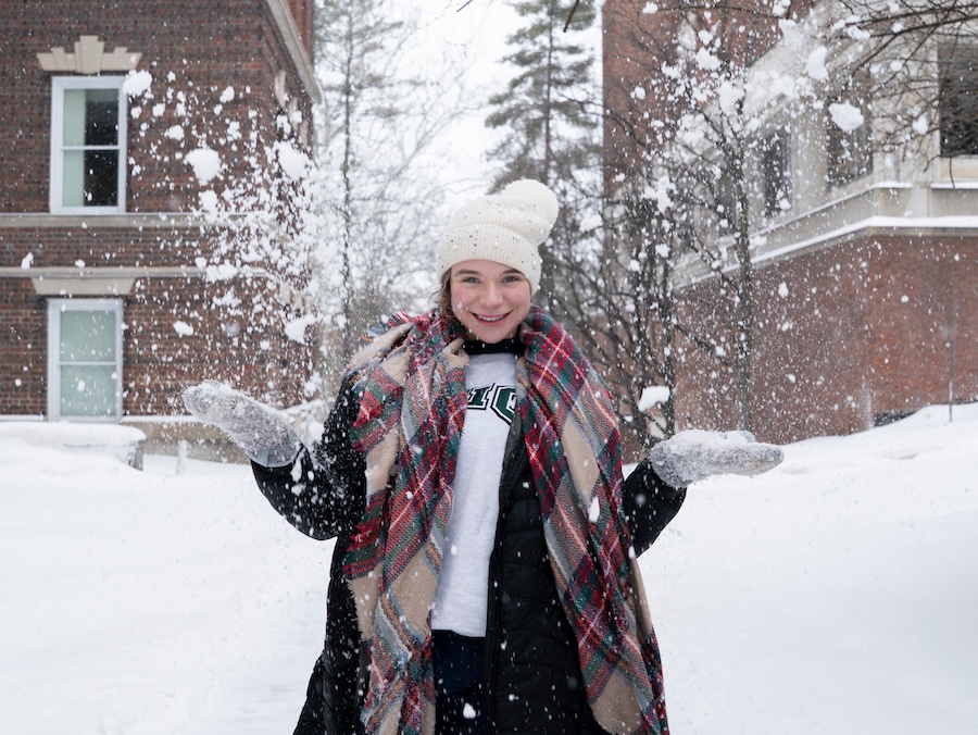A young woman dressed in wintry clothes smiles at the camera and tosses snow into the air with both hands