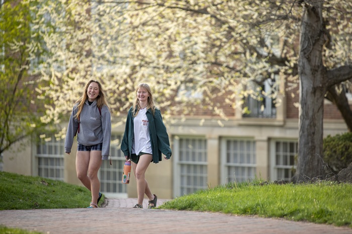 Two students walk on a brick path on the College Green while colorful trees are in the background