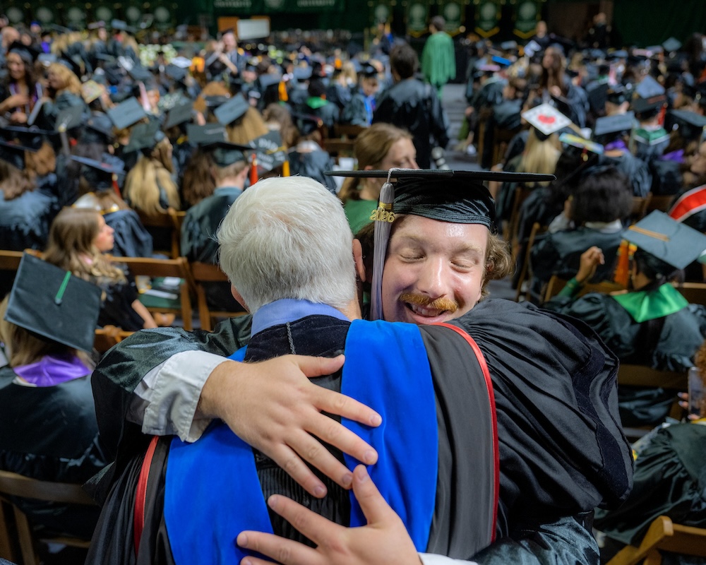 a student wearing a cap and gown hugs a faculty member at the 2026 Spring Commencement ceremony