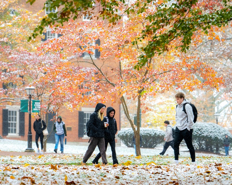 OHIO students walk on the College Green, where there is snow on the ground and in the trees, and coloful Fall leaves on the trees and also on the ground.