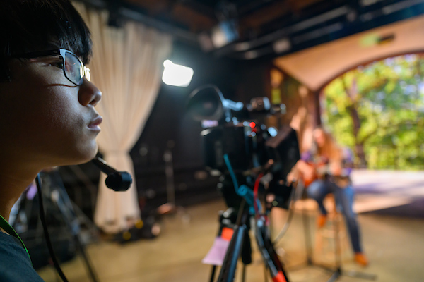 A camp participant wearing a headset records video of a person playing guitar on the sound stage.