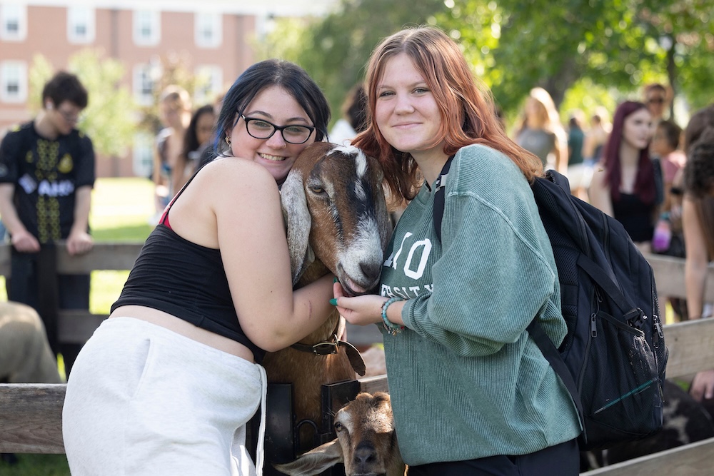 two students pose with a goat at a petting zoo event 