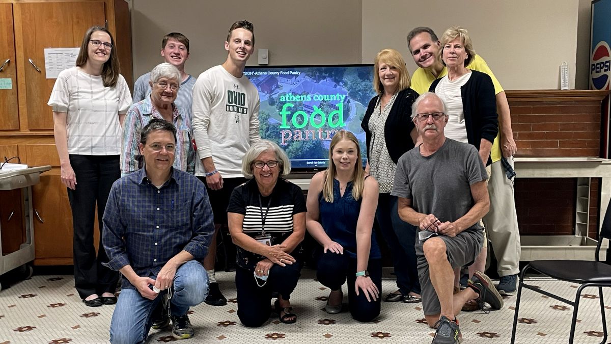 Jack Cowman and members of the Athens Food Pantry pose for a photo before a TV screen.