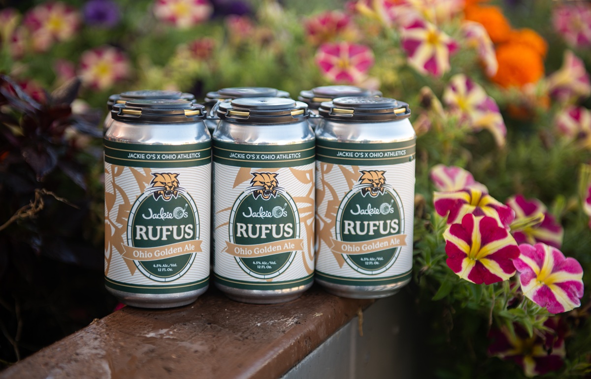 A six-pack of Jackie O's Rufus Ohio Golden Ale is shown near colorful flowers