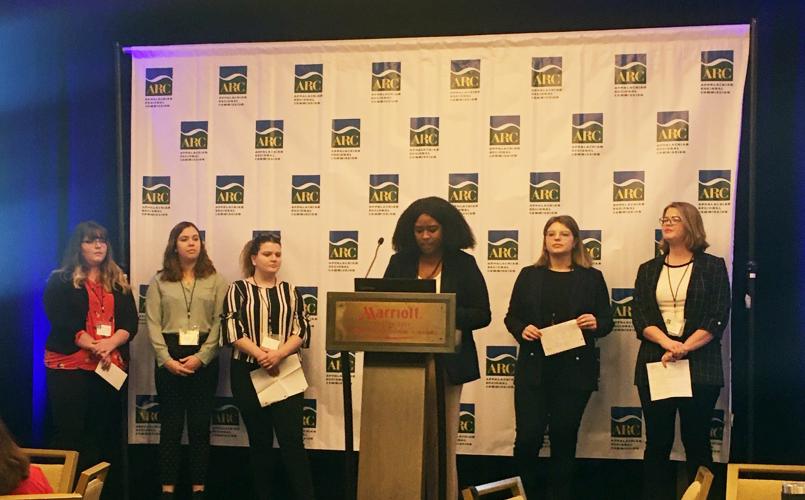 Six students stand on stage, with one at the podium while presenting