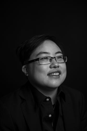 Black and white image of a young East Asian person with glasses smiling and laughing, looking slightly away from the camera