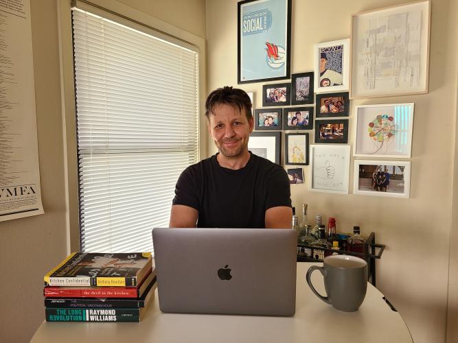 Denis Flaschner sits behind a desk with a stack of books, Apple laptop, mug, with a window and wall of pictures and artwork behind him