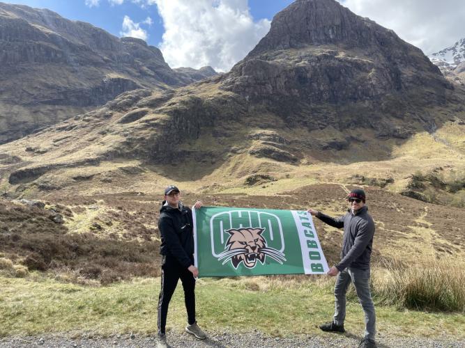 Two people hold up an Ohio University athletics flag in front of a hilly landscape