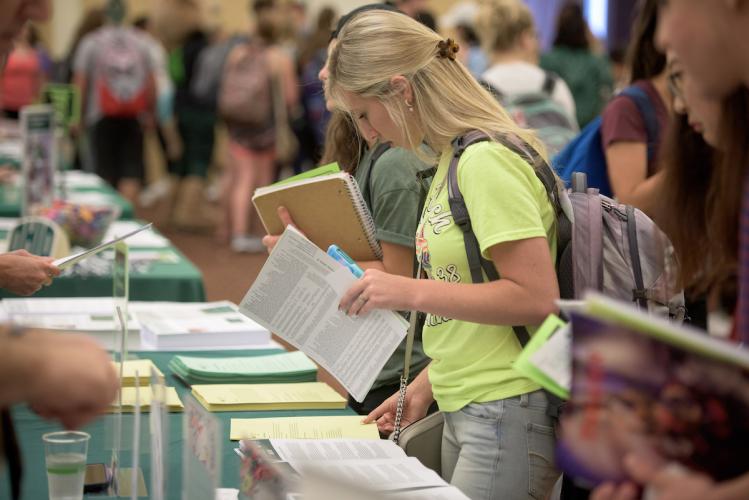 A student looking at flyers at a table while students walk around behind her, looking at other displays