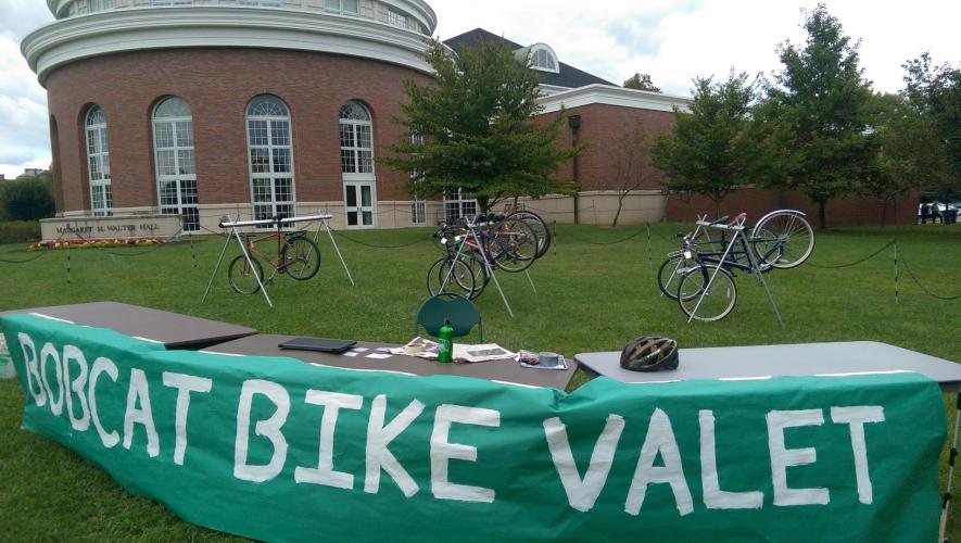 A table set up with a green cloth hanging over it that says "BOBCAT BIKE  VALET" with several bikes on the lawn behind it, with Walter Hall in the background
