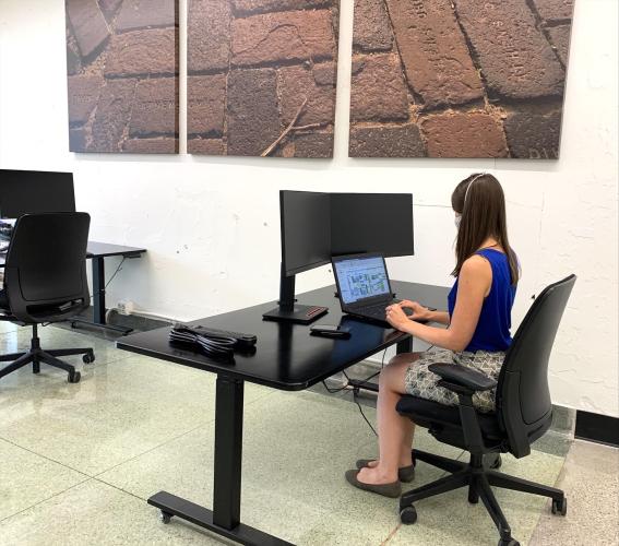 A lady sits at a desk using her laptop; the desk has a monitor and keyboard, and a similar setup is to the right and front. 