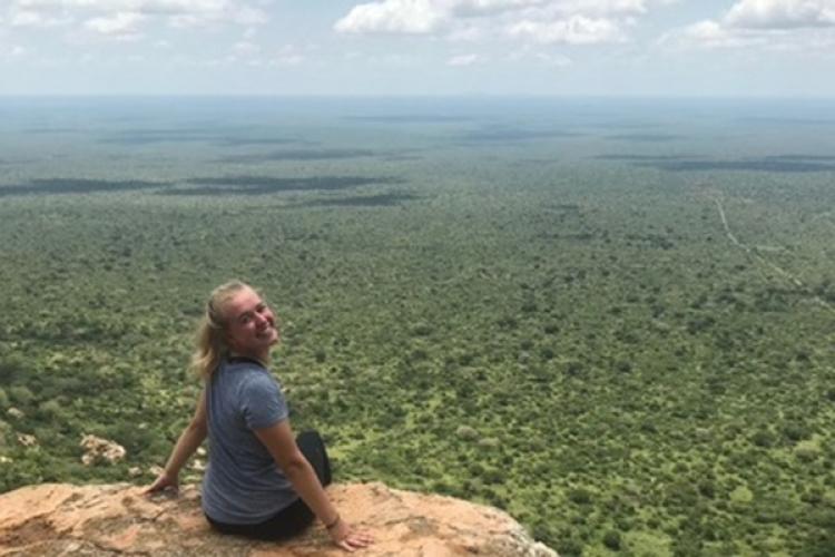 A woman sits on a cliff overlooking an array of trees, with her face turned towards the camera with a smile