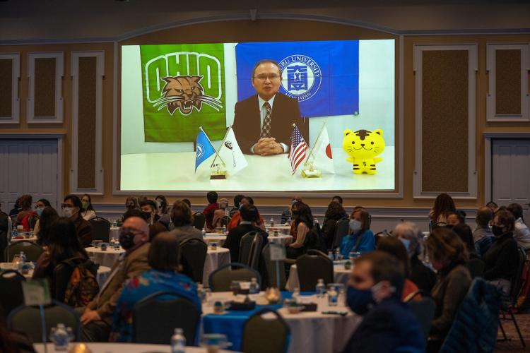 Chubu University Professor Nobuyuki Kawauchi giving an acceptance speech broadcasted on a screen while onlookers watch from dining tables