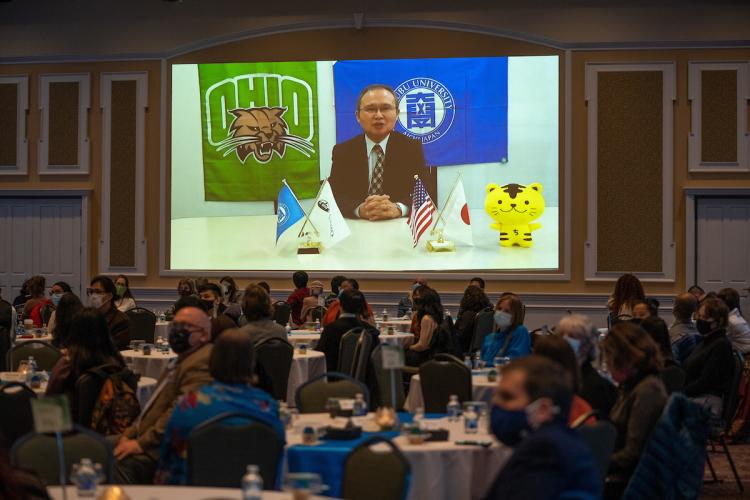 Chubu University Professor Nobuyuki Kawauchi giving an acceptance speech broadcasted on a screen while onlookers watch from dining tables