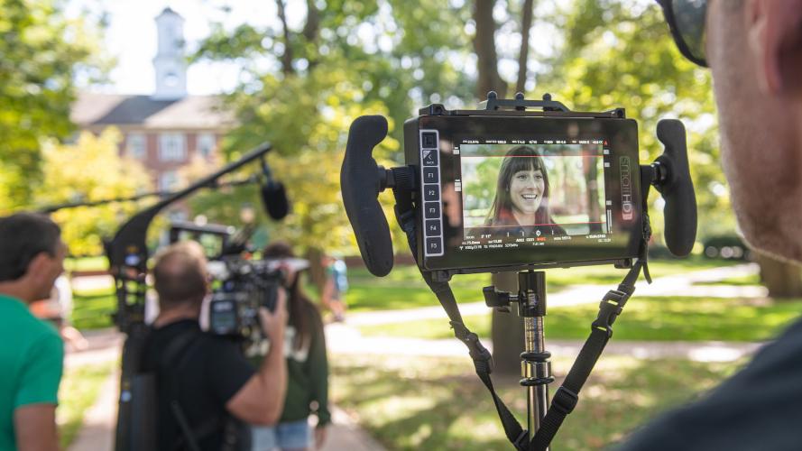 A camera operator is holding a camera screen on College Green, with the screen focused on a smiling student