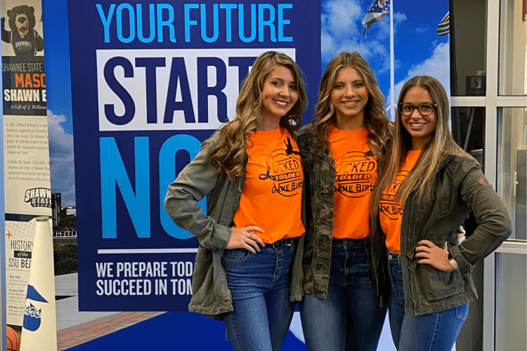 Three smiling high school students pose in front of a poster