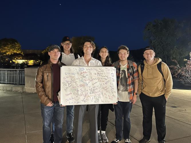 Students stand together holding a large white board with writing all over it in a parking lot