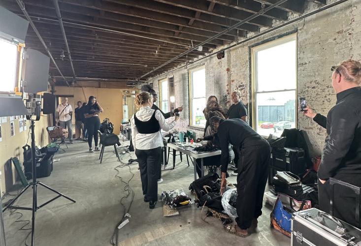 Two Ohio University students stand in front of a table interviewing people behind it, with the room cluttered in lights, boxes and things to set up for a fashion photoshoot