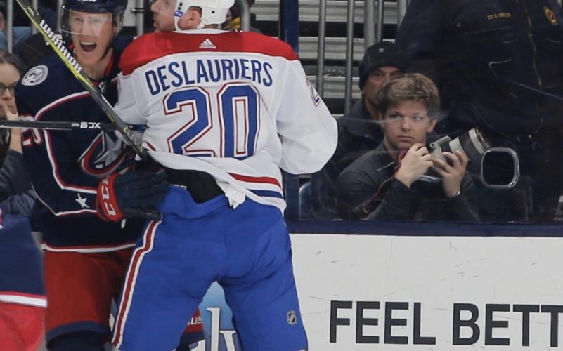 Two hockey players collide in the foreground as a photographer observes from the stands