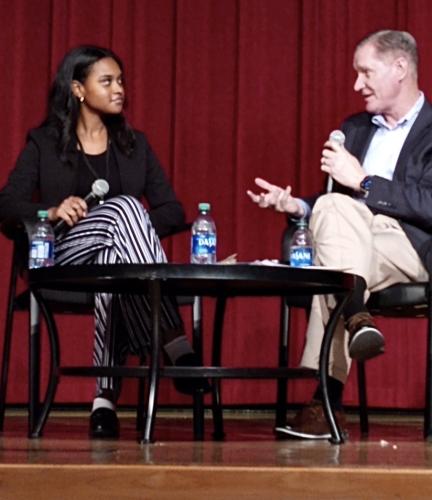 Two people sit side-by-side on a stage, both holding a microphone. The man on the right is speaking directly to the woman on the left