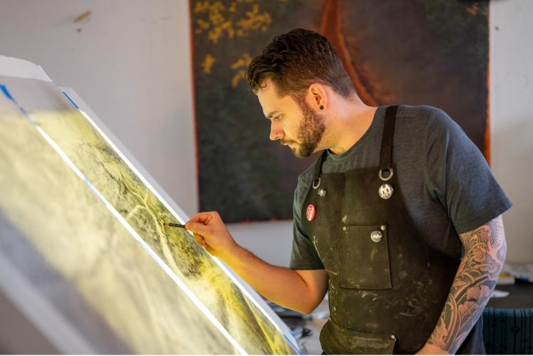 An artist stands at an illuminated easel, working on a print